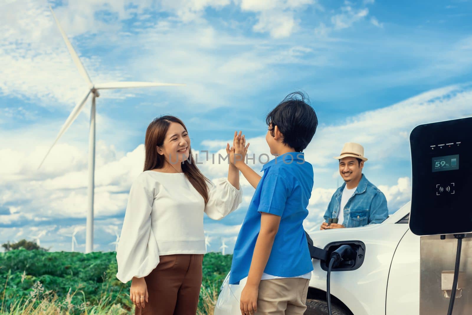 Concept of progressive happy family enjoying their time at wind farm with electric vehicle. Electric vehicle driven by clean renewable energy from wind turbine generator for charging station.