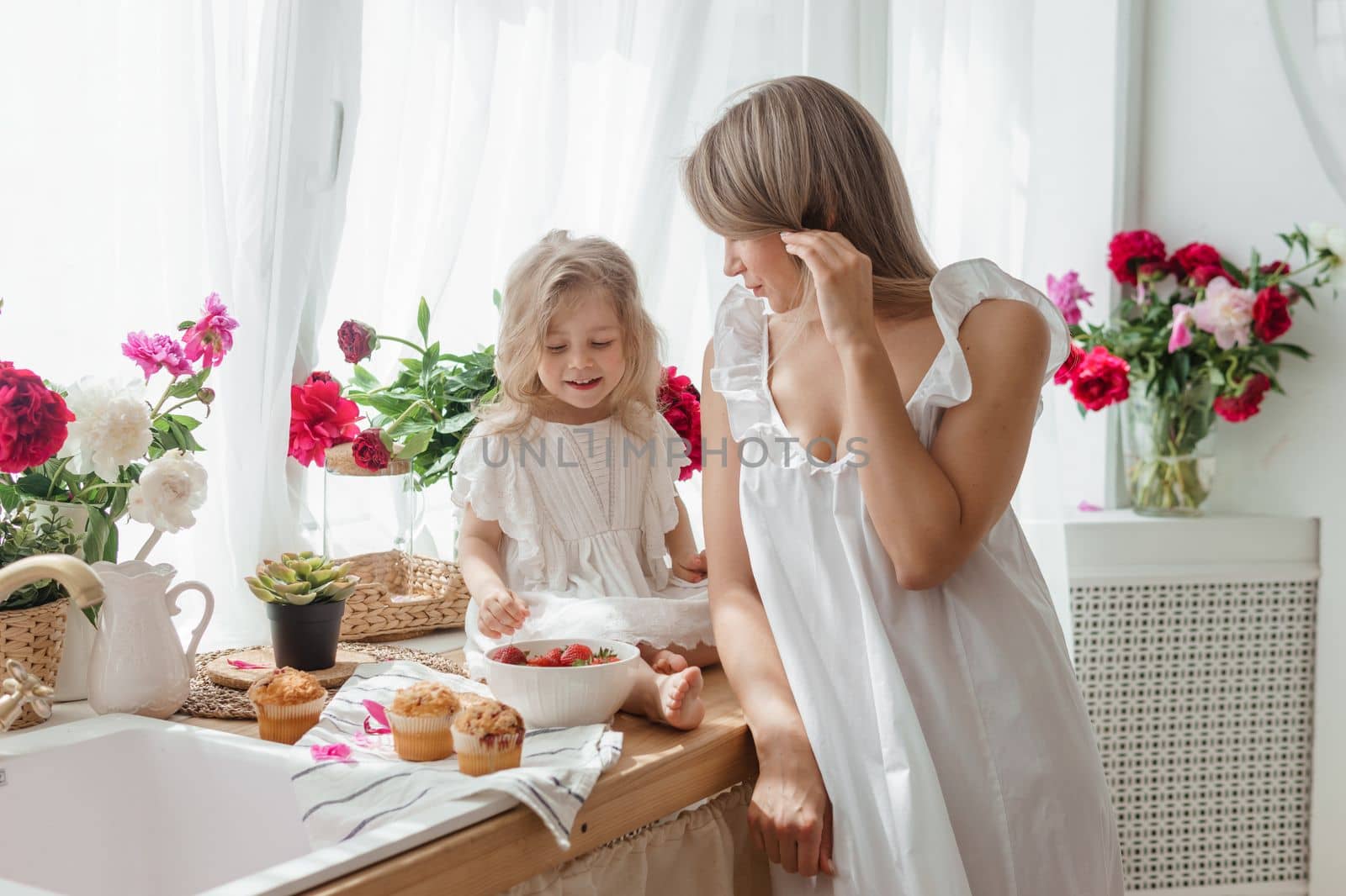 A little blonde girl with her mom on a kitchen countertop decorated with peonies. The concept of the relationship between mother and daughter. Spring atmosphere. by Annu1tochka