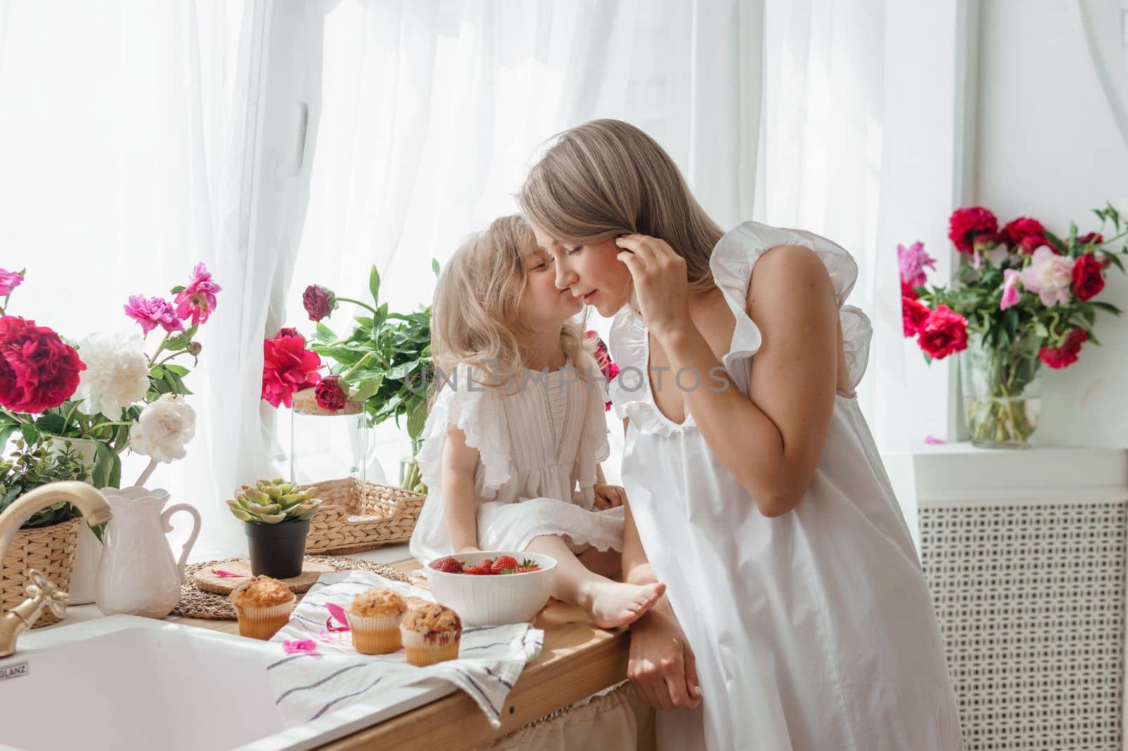 A little blonde girl with her mom on a kitchen countertop decorated with peonies. The concept of the relationship between mother and daughter. Spring atmosphere.