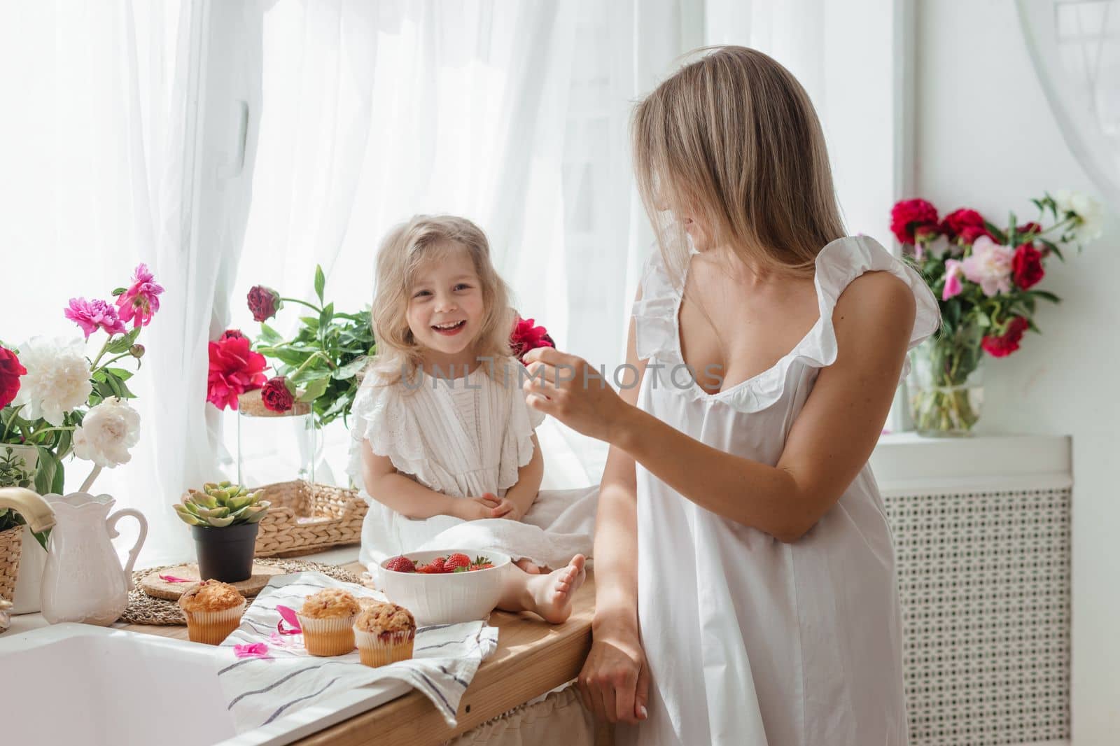 A little blonde girl with her mom on a kitchen countertop decorated with peonies. The concept of the relationship between mother and daughter. Spring atmosphere.