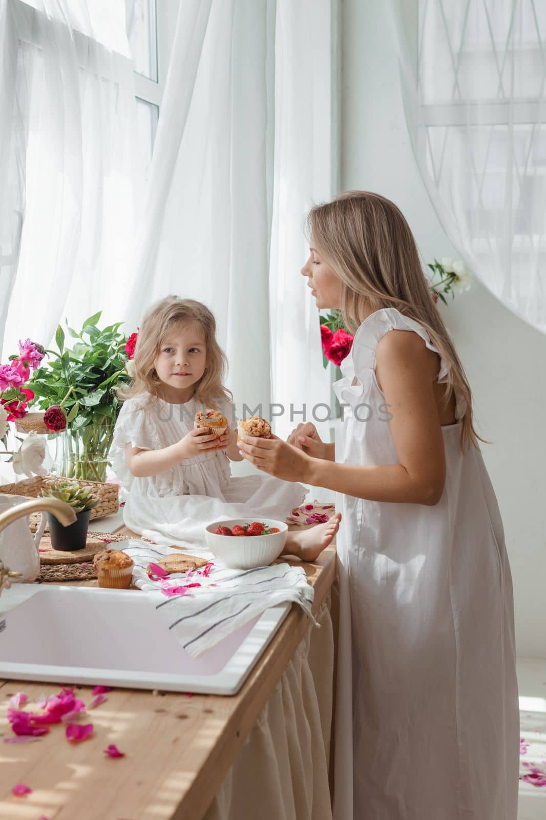 A little blonde girl with her mom on a kitchen countertop decorated with peonies. The concept of the relationship between mother and daughter. Spring atmosphere.