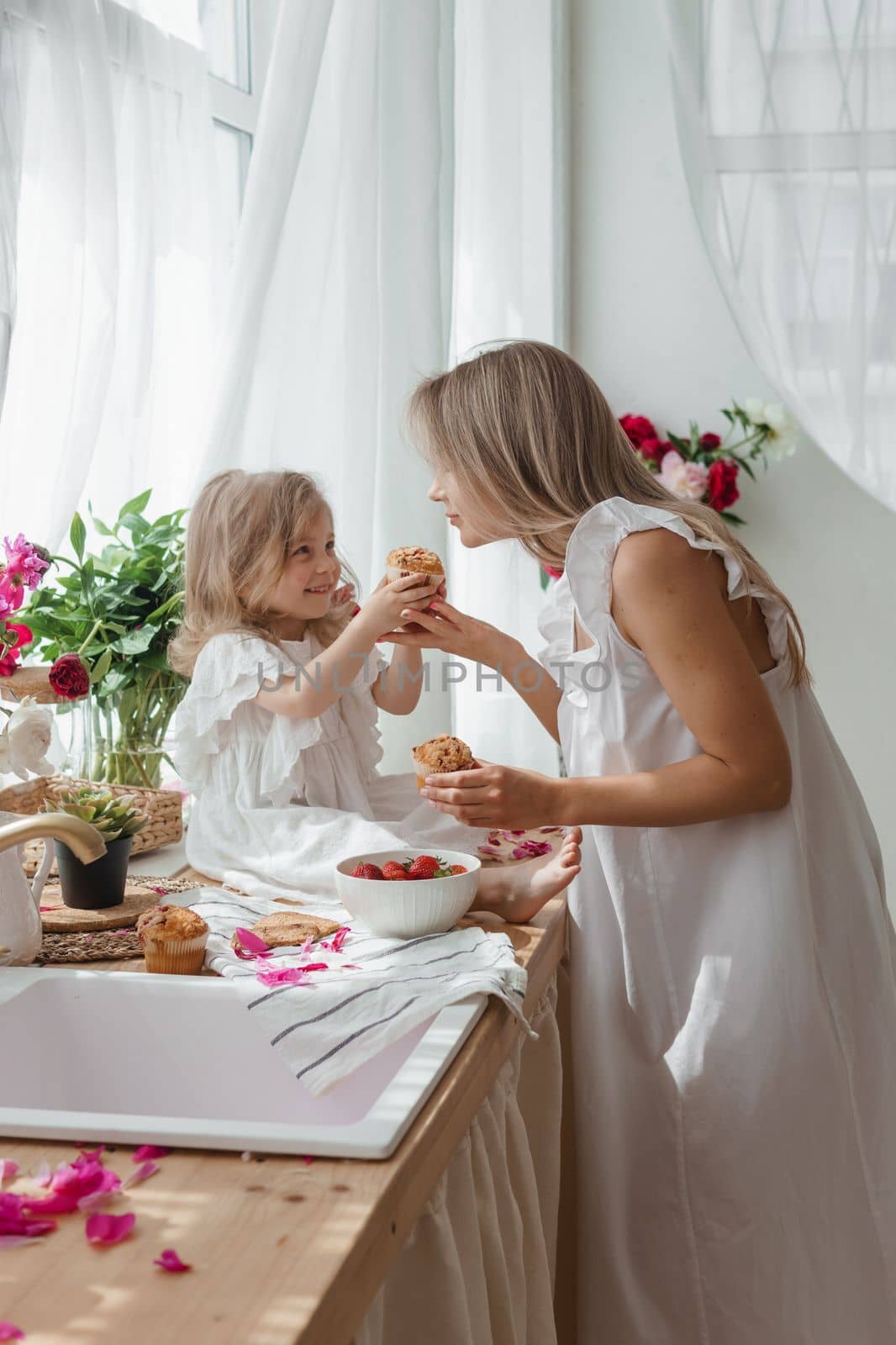 A little blonde girl with her mom on a kitchen countertop decorated with peonies. The concept of the relationship between mother and daughter. Spring atmosphere.