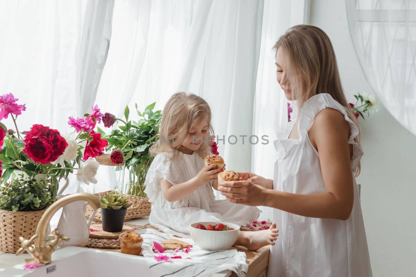 A little blonde girl with her mom on a kitchen countertop decorated with peonies. The concept of the relationship between mother and daughter. Spring atmosphere.