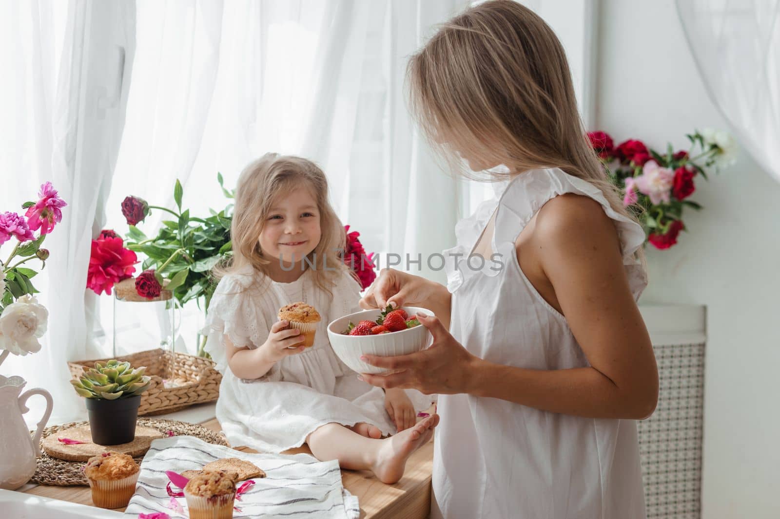 A little blonde girl with her mom on a kitchen countertop decorated with peonies. The concept of the relationship between mother and daughter. Spring atmosphere. by Annu1tochka