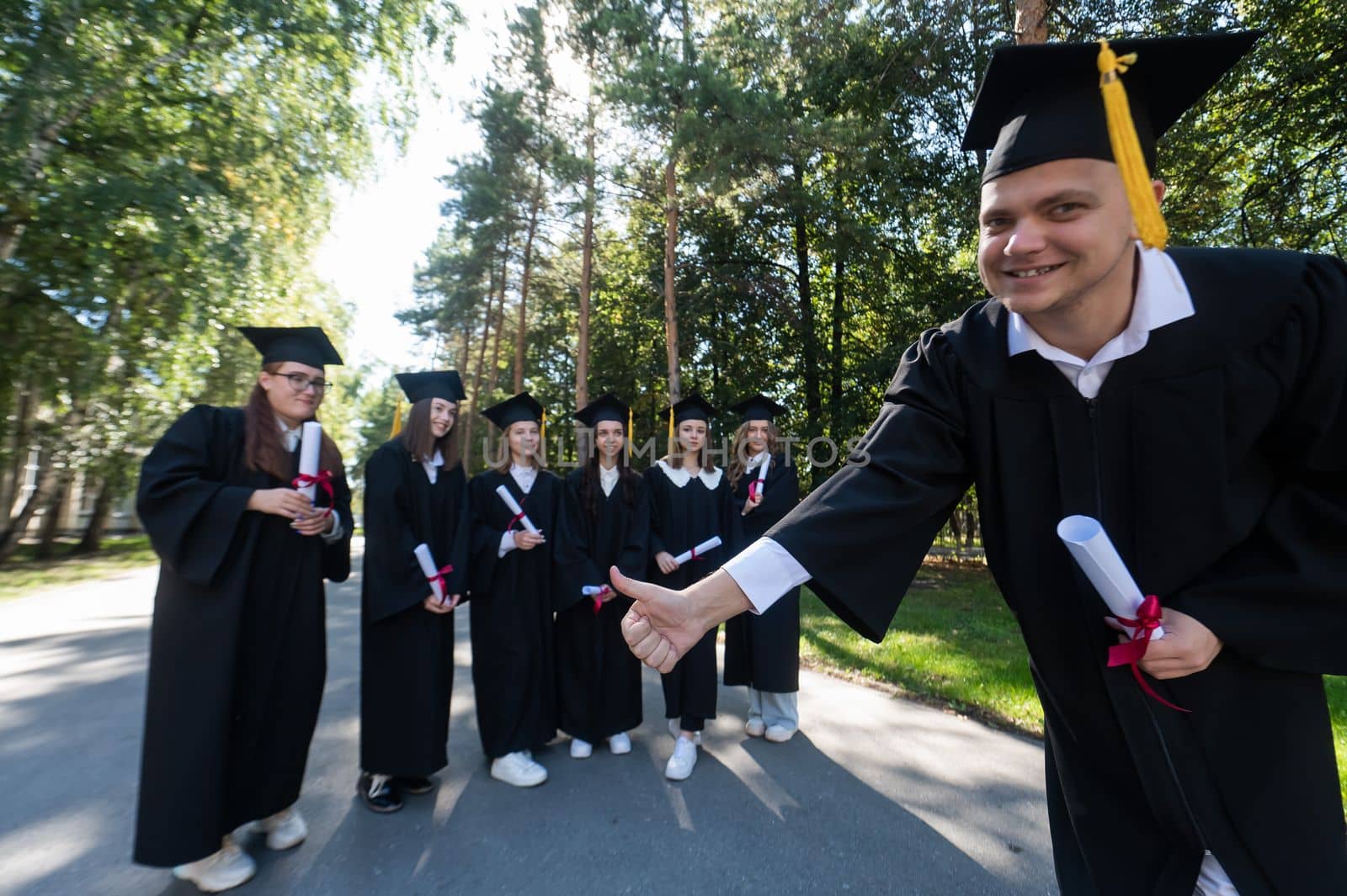 Happy young caucasian male graduate showing thumbs up. A group of graduate students outdoors