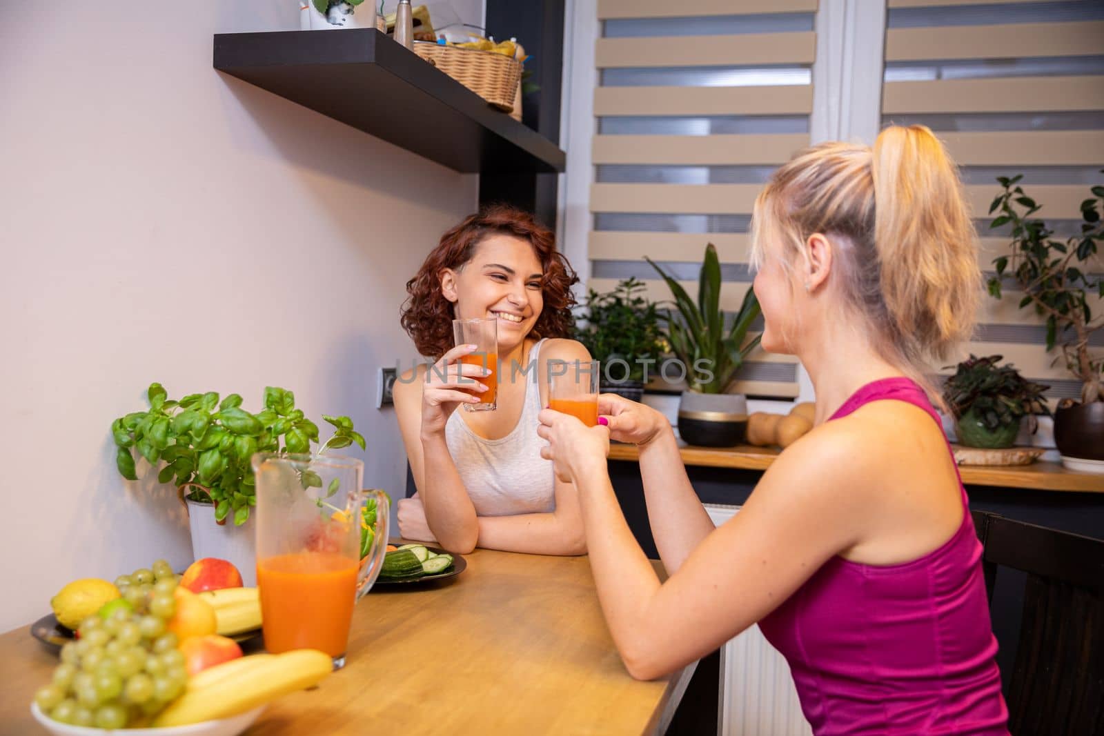 two women are talking in the kitchen sitting across from each other and smiling. One woman can be seen in profile and the other is sitting against a curtained window. on the table are plates with fruits and vegetables.