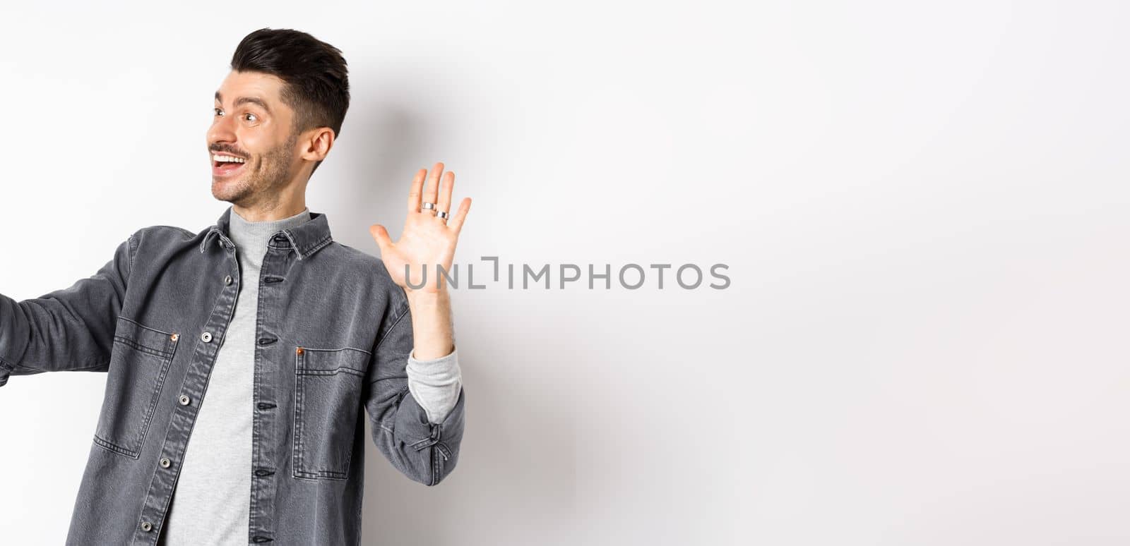 Young man video chatting on mobile phone, waiving hand at smartphone camera, greeting friend online, standing on white background by Benzoix