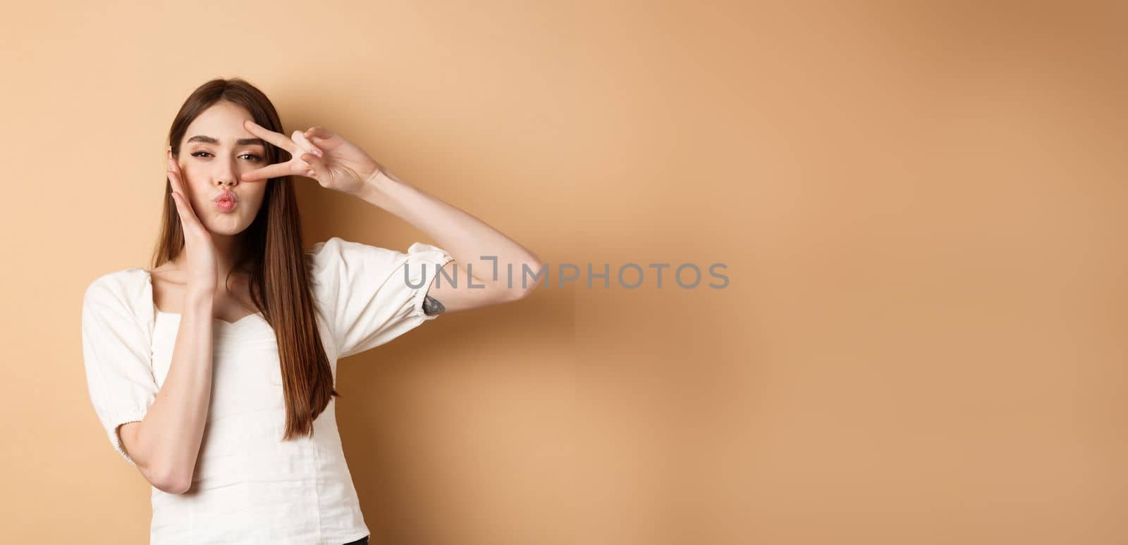 Cute woman showing v-sign and pucker lips for kiss, touching face with silly expression, standing on beige background.