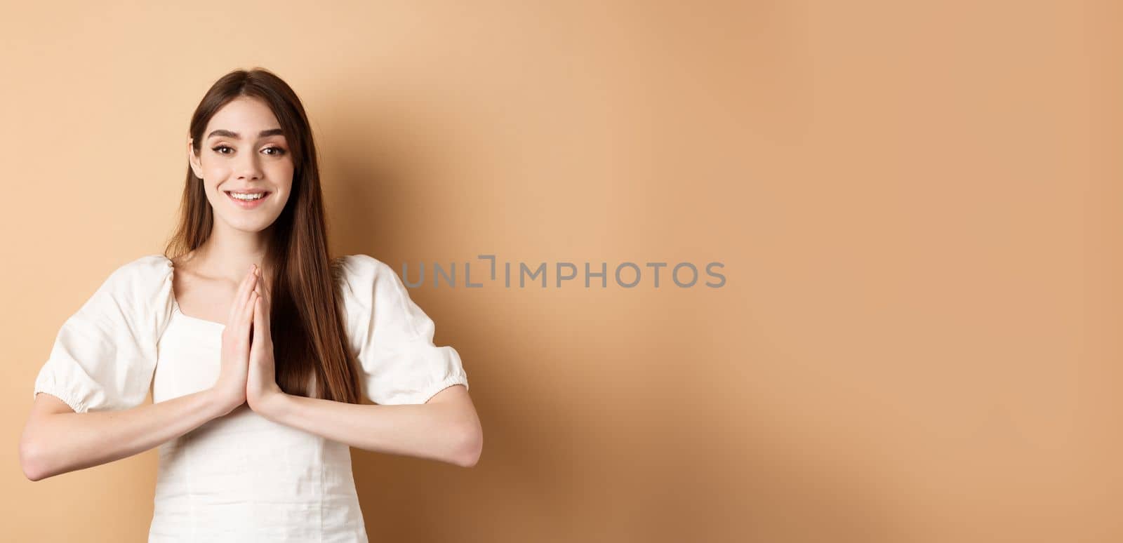 Pretty please. Cute young woman asking for favour, holding hands in begging pose and smiling, need help, standing on beige background by Benzoix