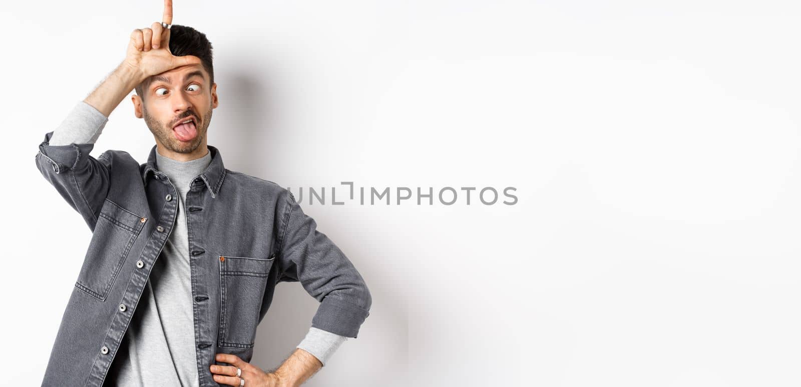 Funny young man making faces and showing tongue with loser sign on forehead, squinting eyes, standing on white background by Benzoix