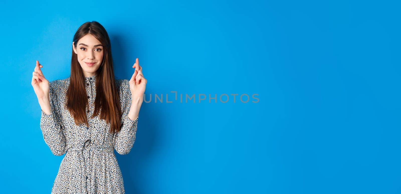 Hopeful smiling woman cross fingers for good luck and looking positive, praying or making wish, standing on blue background by Benzoix