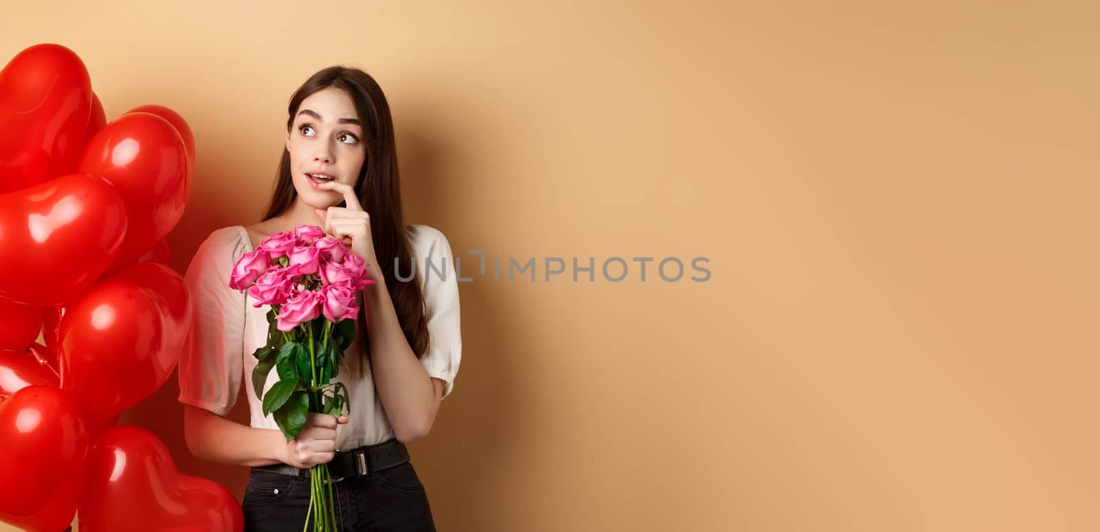 Dreamy young woman holding bouquet of roses and thinking about secret admirer on Valentines day, looking at upper left corner and biting finger, standing near red romantic balloons.