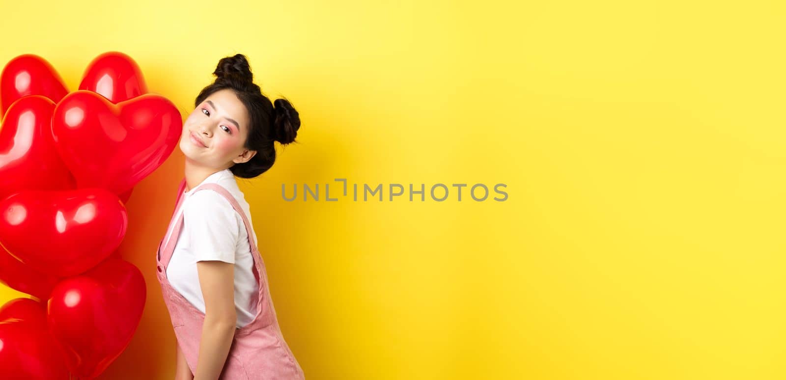 Valentines day concept. Stylish teenage asian girl posing near red hearts balloons, wear outfit for romantic date, standing happy on yellow background.