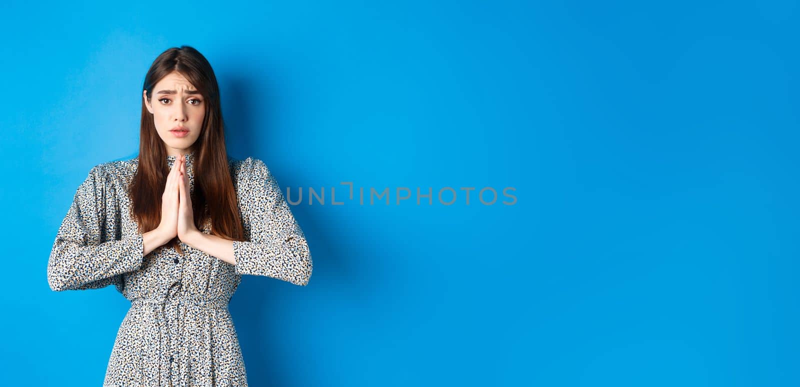 Sad girl asking for help, begging you with troubled face expression, standing in dress on blue background.