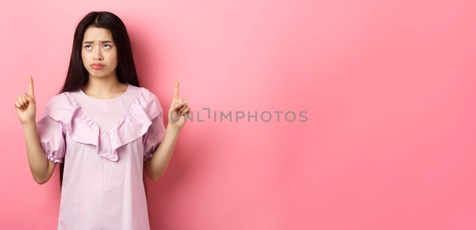 Sad pitiful asian girl sulking, pointing and looking up with disappointed face, feeling upset, standing against pink background.
