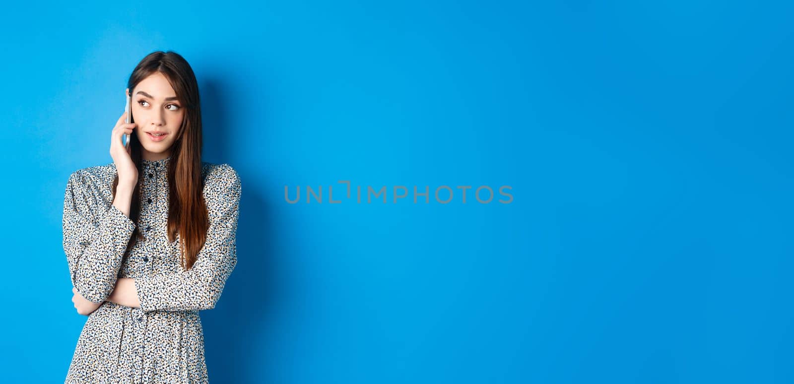 Modern beautiful woman talking on mobile phone, looking aside at logo and making call, standing against blue background by Benzoix