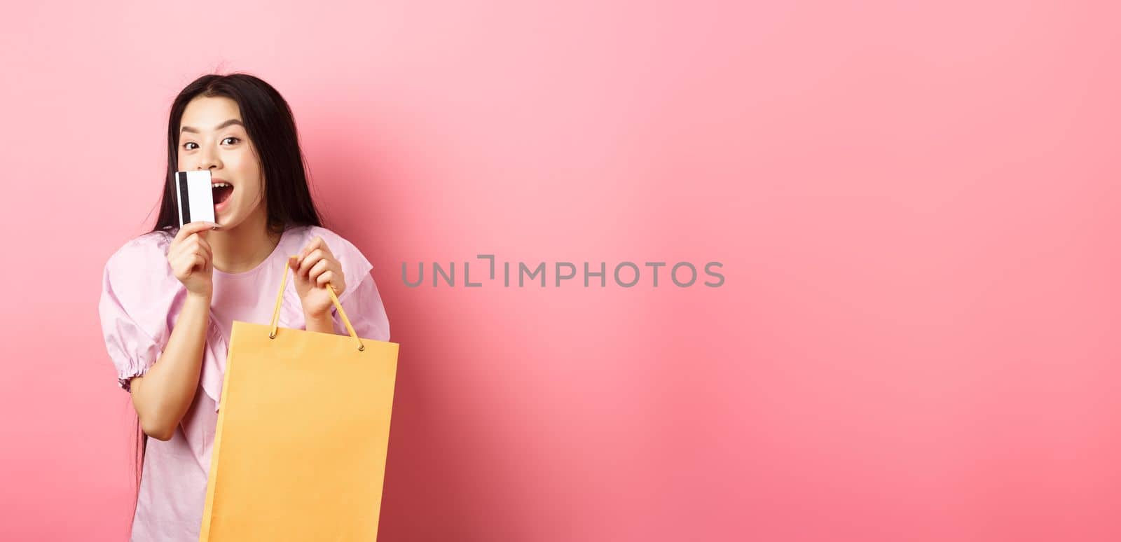 Shopping. Excited beautiful asian woman buying in stores, holding paper bag and kissing plastic credit card, standing against pink background.