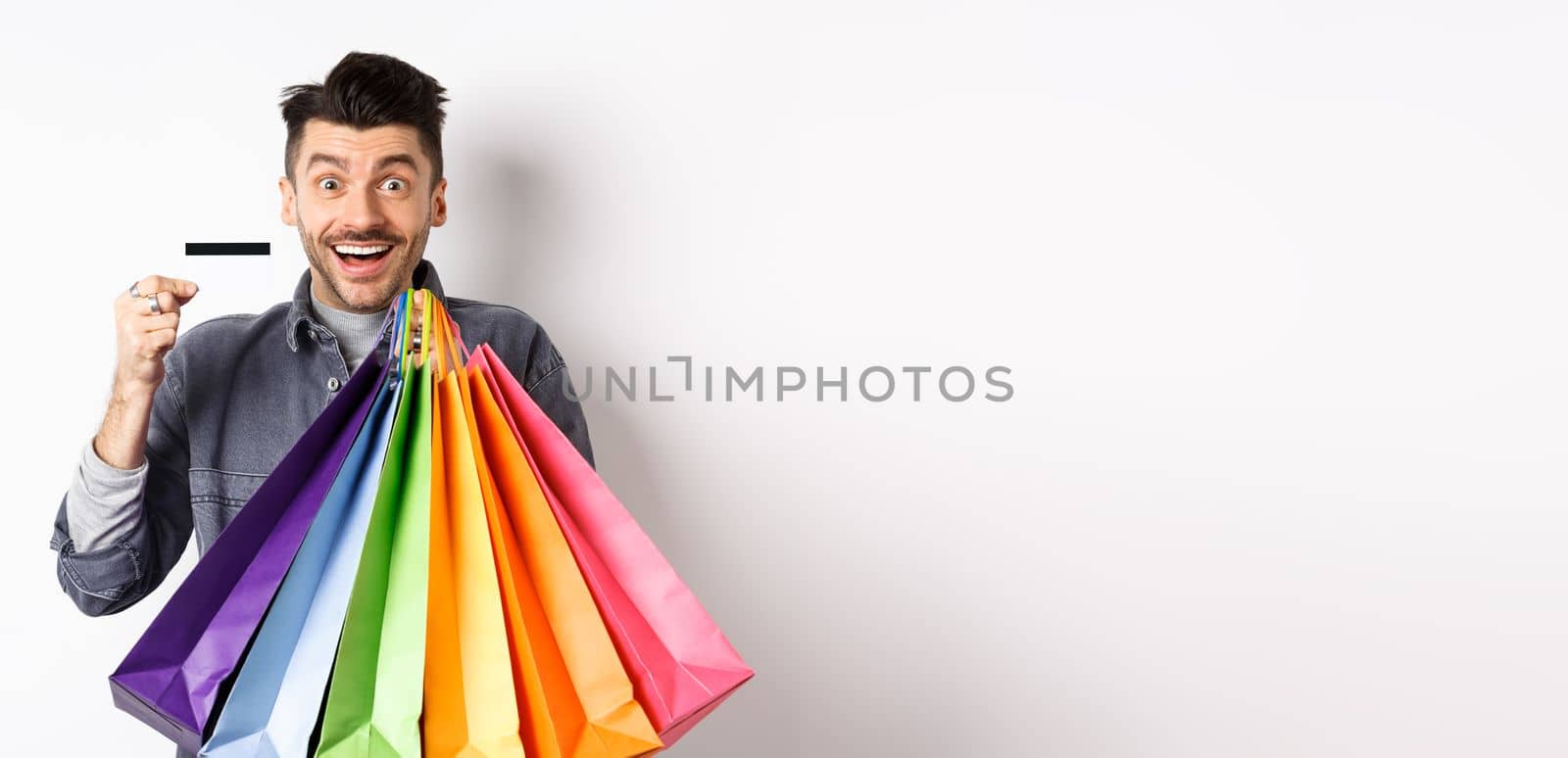 Happy guy shopaholic holding colorful shopping bags and showing plastic credit card, smiling excited, standing white background.