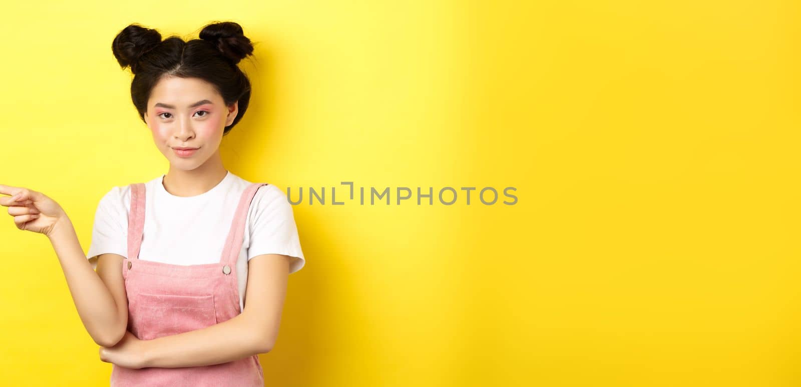 Beautiful teenage asian girl with bright makeup, pointing finger left at banner and smiling, yellow background by Benzoix