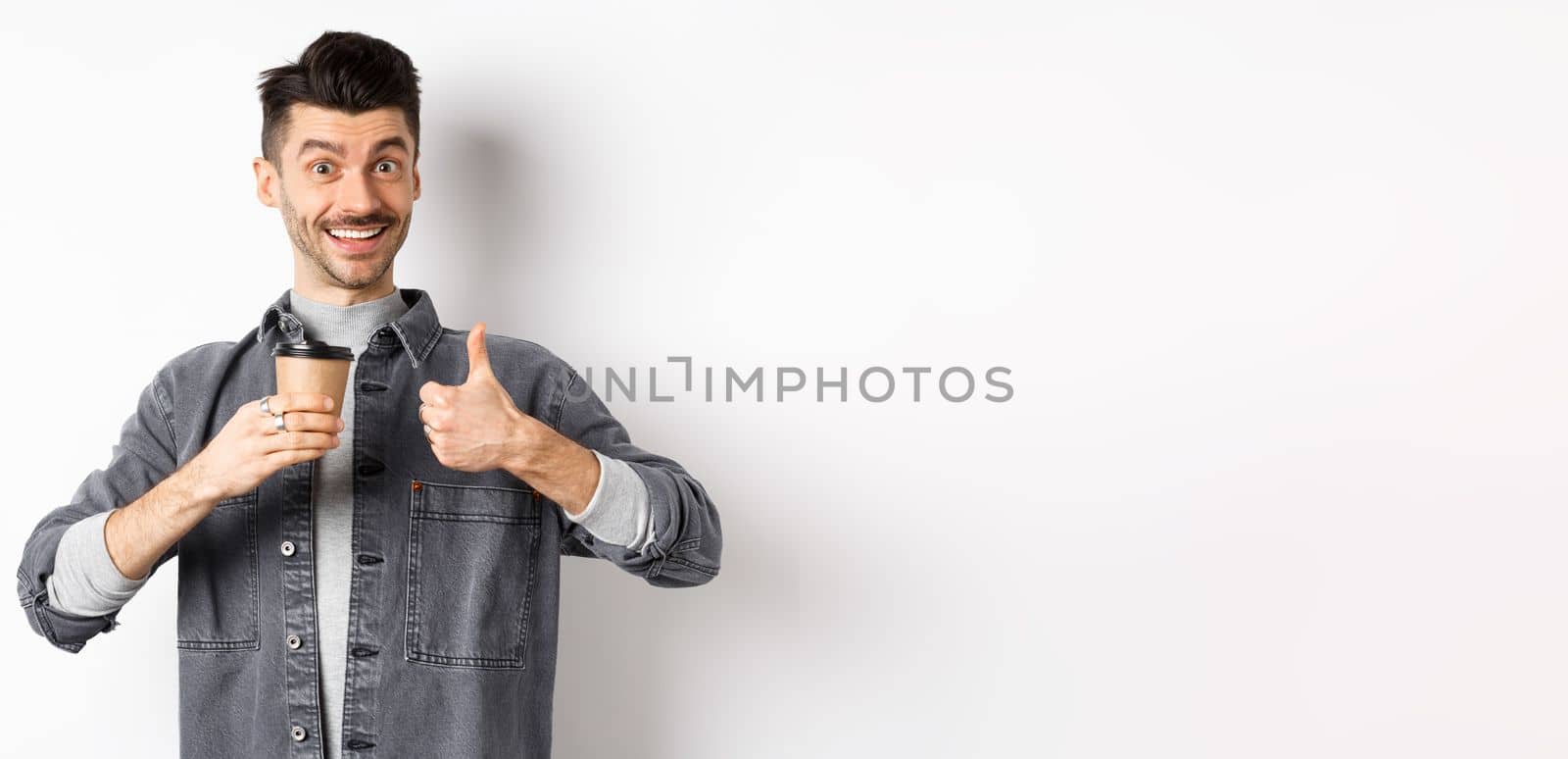 Very good drink. Smiling stylish guy drinking coffee and showing thumbs up, praise coffee shop, standing satisfied on white background.