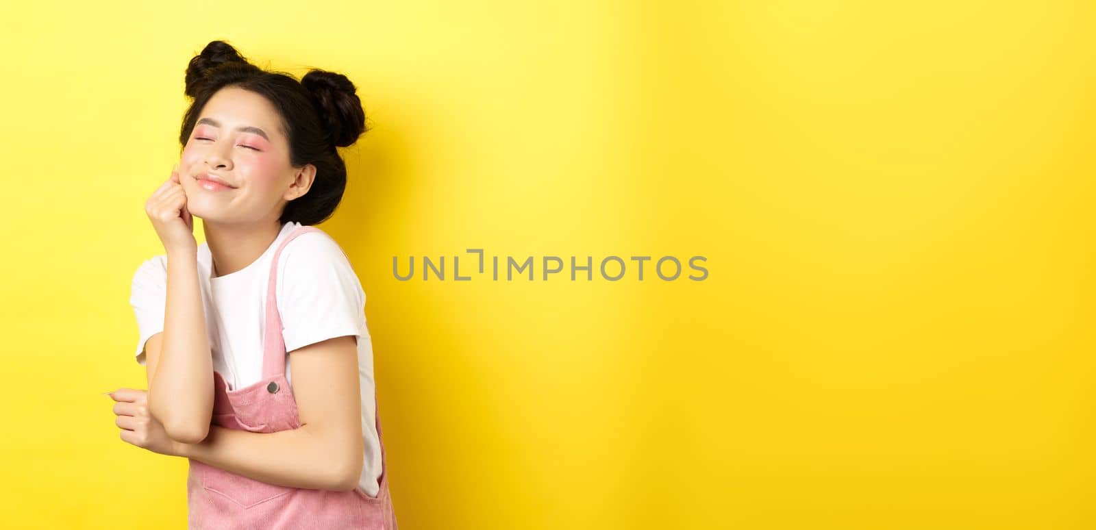 Beautiful happy woman with bright pink makeup, close eyes and touch soft skin, smiling excited, standing in summer clothes on yellow background by Benzoix