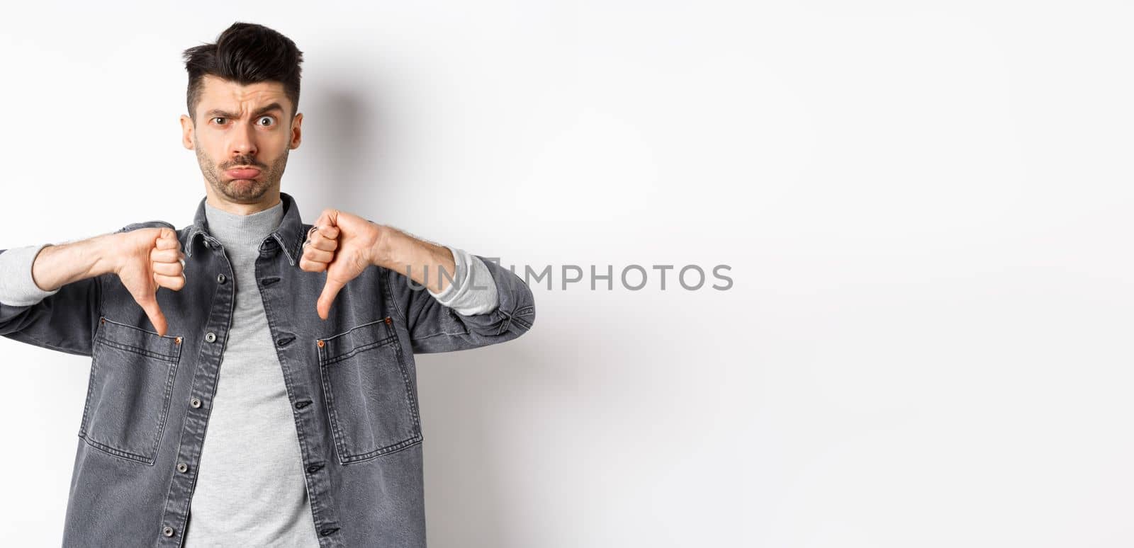 Disappointed funny man frowning and looking upset, showing thumbs down displeased, standing on white background by Benzoix