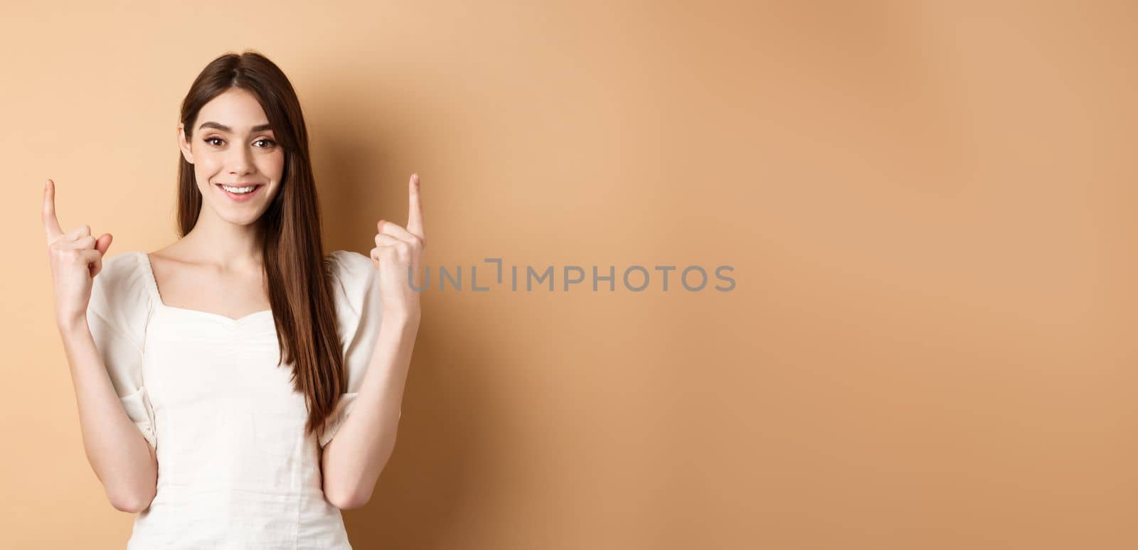 Attractive caucasian woman in dress pointing fingers up, smiling and showing promo banner, standing on beige background.
