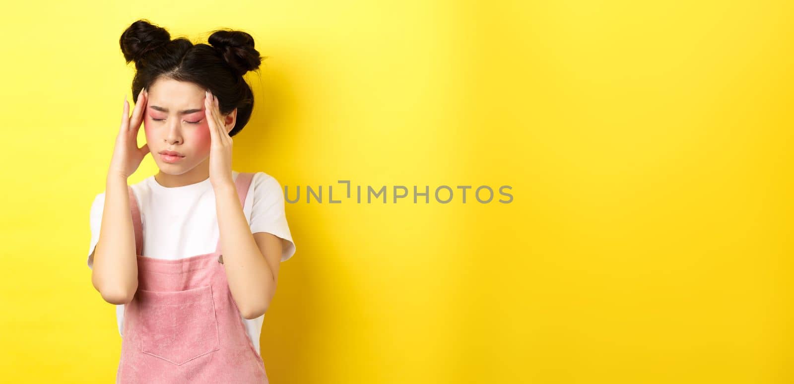 Image of stylish asian woman with bright makeup, having headache, touching head and looking tired, have painful migraine, yellow background.