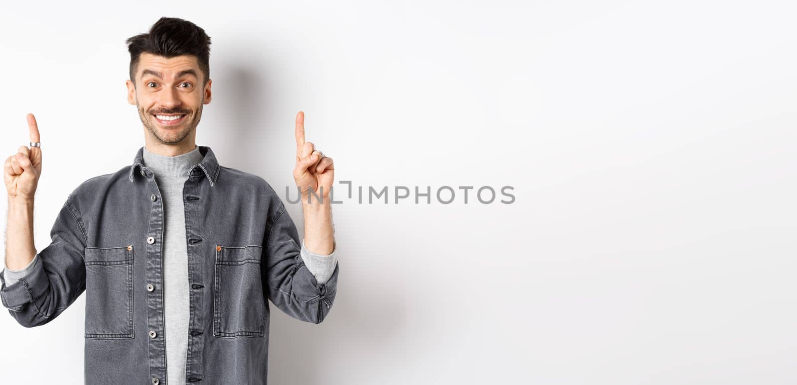 Excited young man smiling and showing advertisement, pointing fingers up at promo offer, standing on white background.
