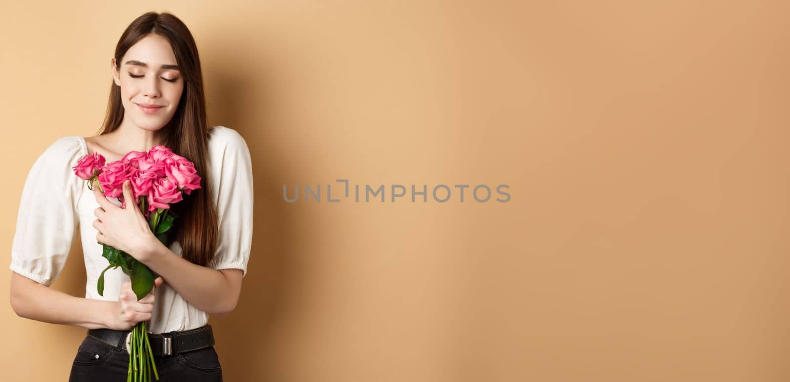 Valentines day. Tender and romantic girl, smell roses and smile with closed eyes. Girlfriend hugging gift flowers from lover, standing on beige background.