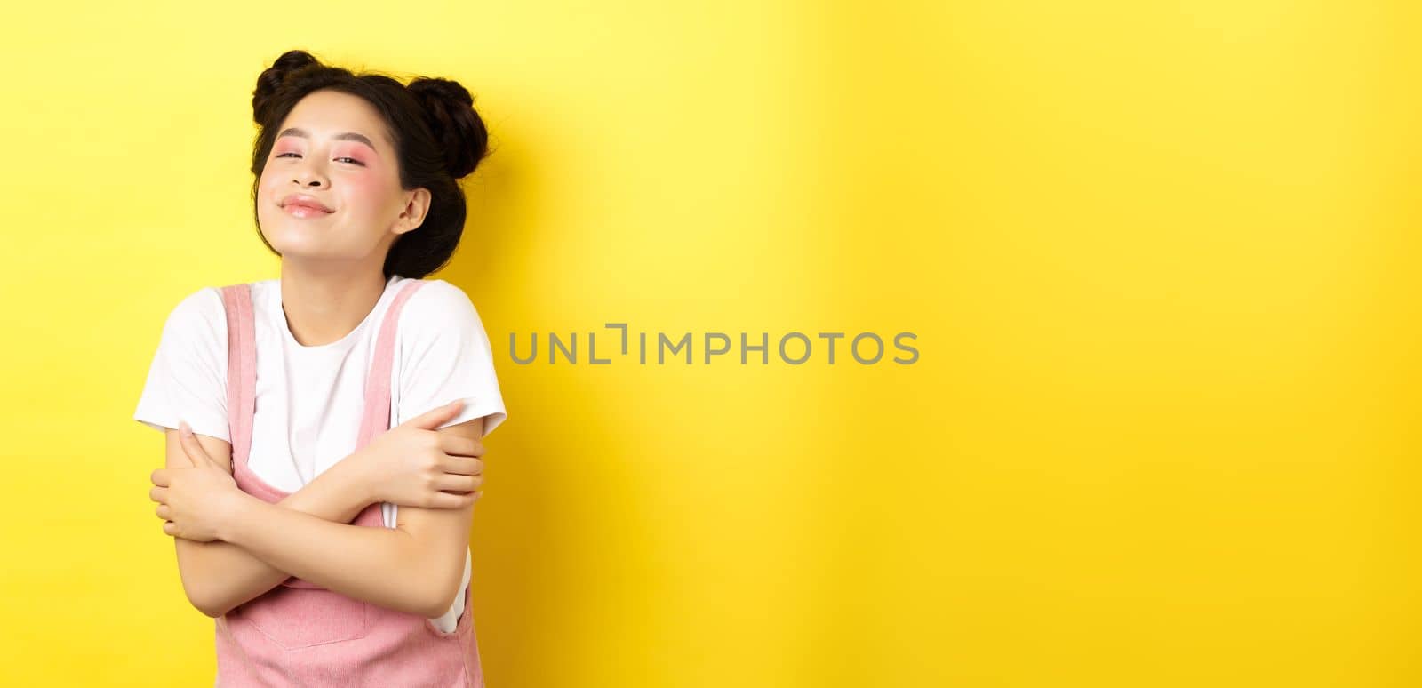 Summer lifestyle concept. Beautiful asian woman hugging herself and smiling romantic, embracing own body, standing with makeup on yellow background.