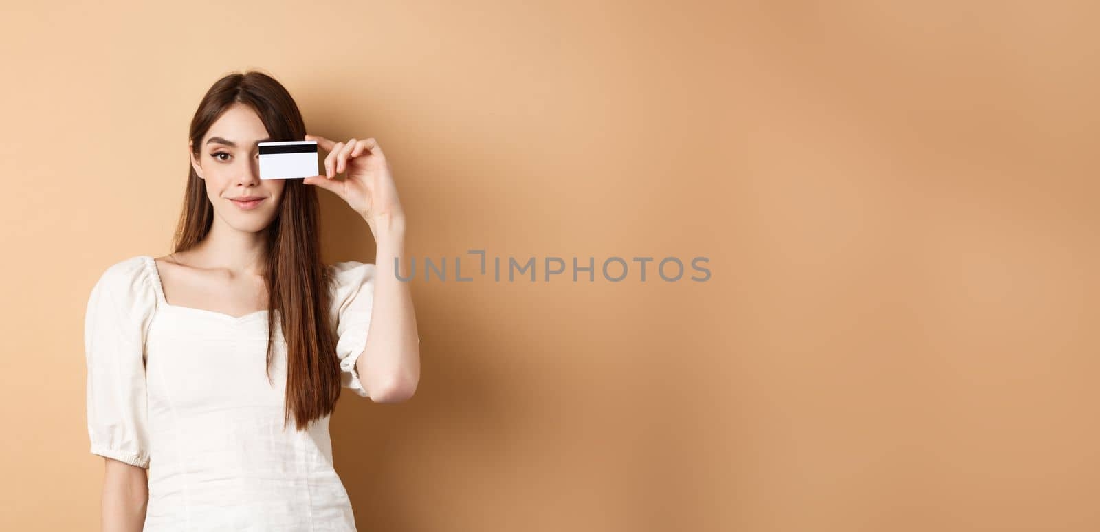 Smiling pretty girl showing plastic credit card over eye and looking satisfied, standing on beige background. Concept of shopping by Benzoix