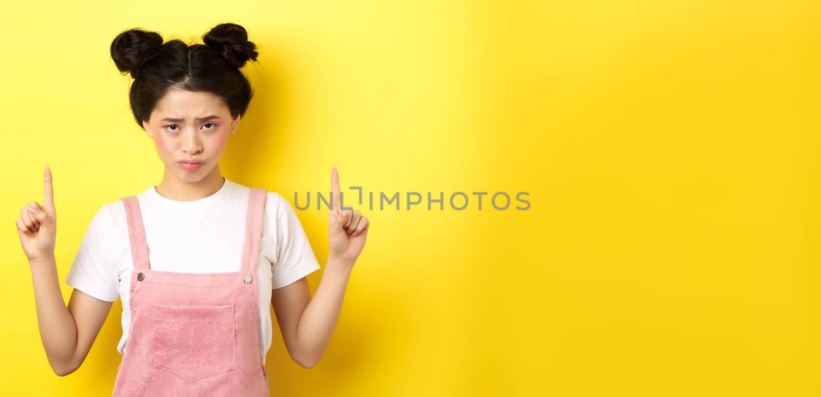 Sad and gloomy asian woman frowning, pointing fingers up and complaining on unfair thing, standing upset on yellow background.