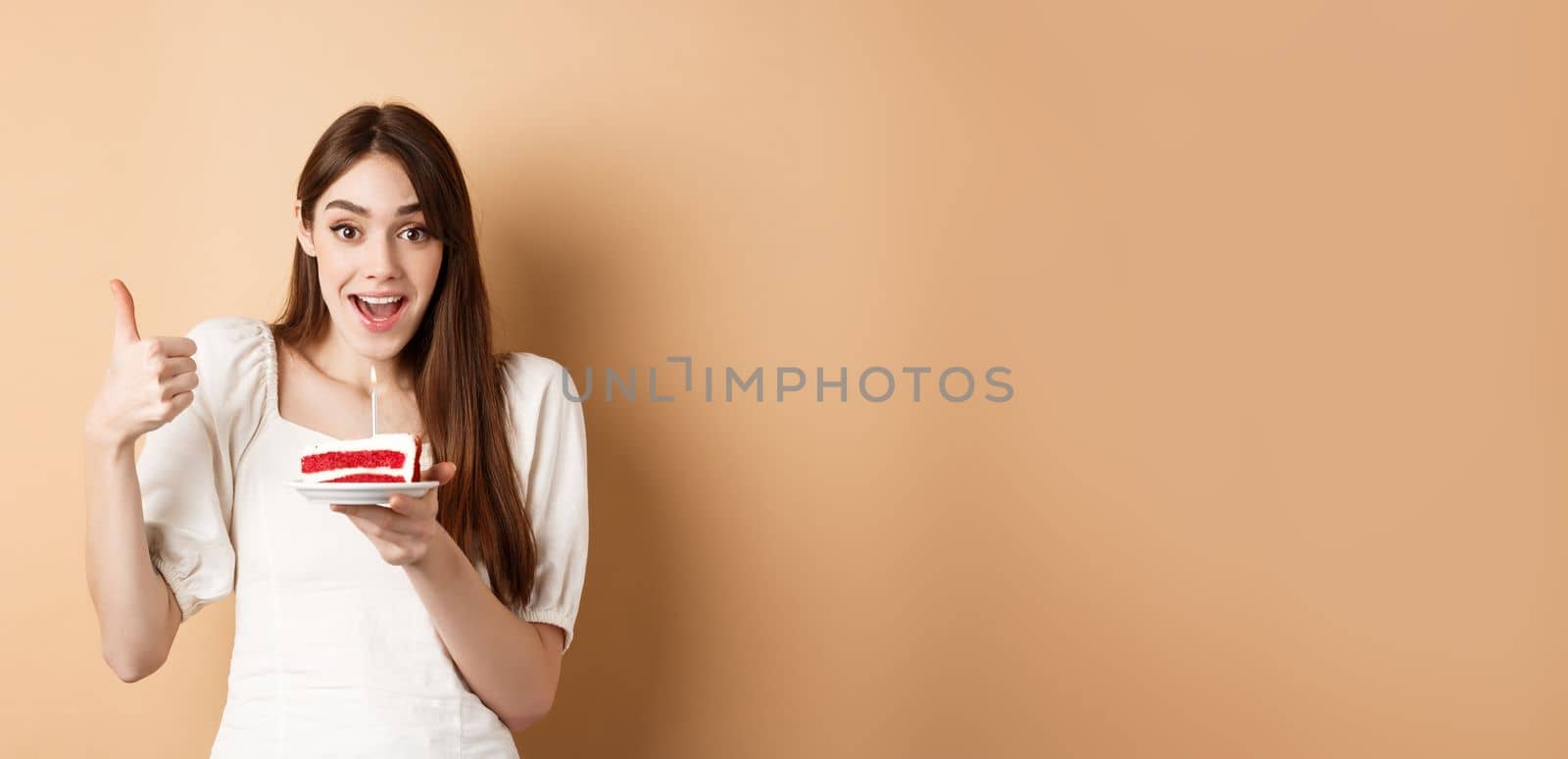 Excited birthday girl show thumb up and hold cake with candle, making wish, satisfied with bday party, smiling happy at camera, beige background.