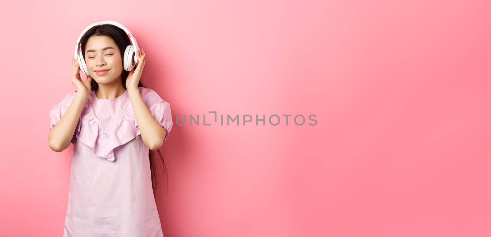 Beautiful asian girl close eyes while listening music in headphones, enjoying soft sound, standing against pink background by Benzoix