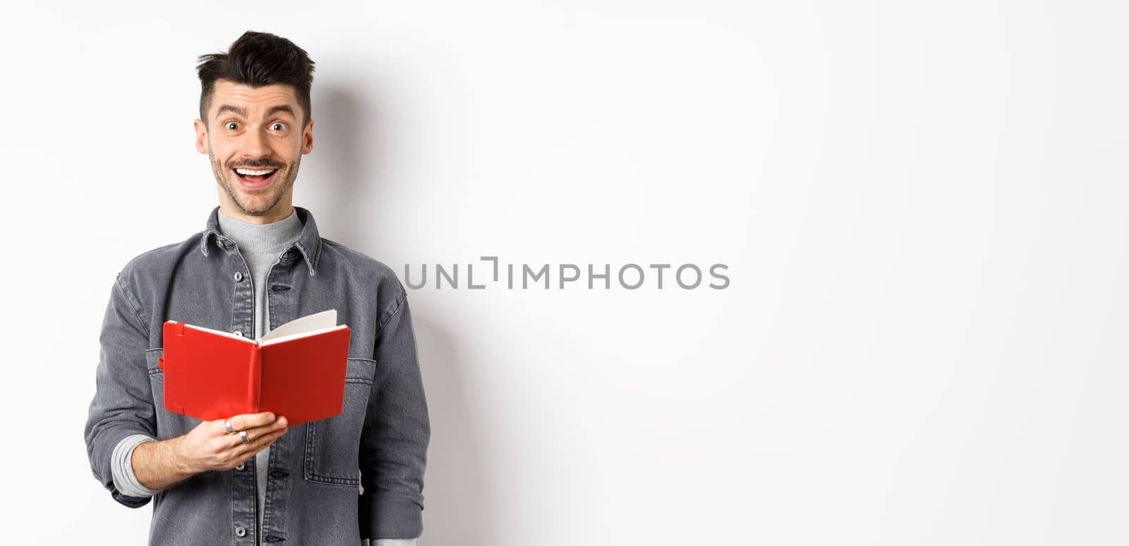 Hobbies and leisure. Happy young man reading planner, holding diary or red journal and smiling, making notes, standing on white background by Benzoix