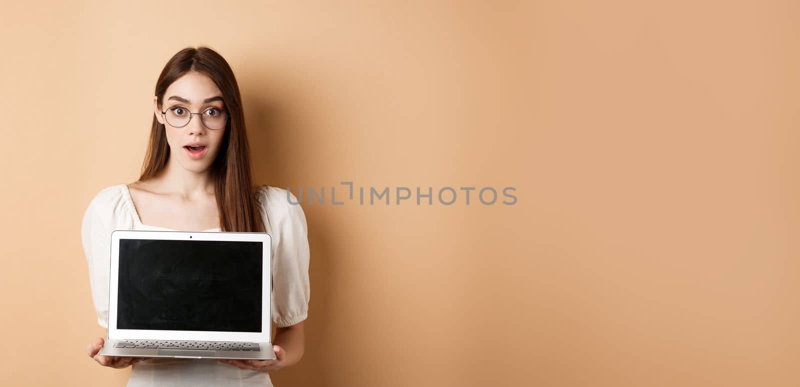 Surprised young woman in glasses showing laptop screen with amazed face, standing on beige background by Benzoix
