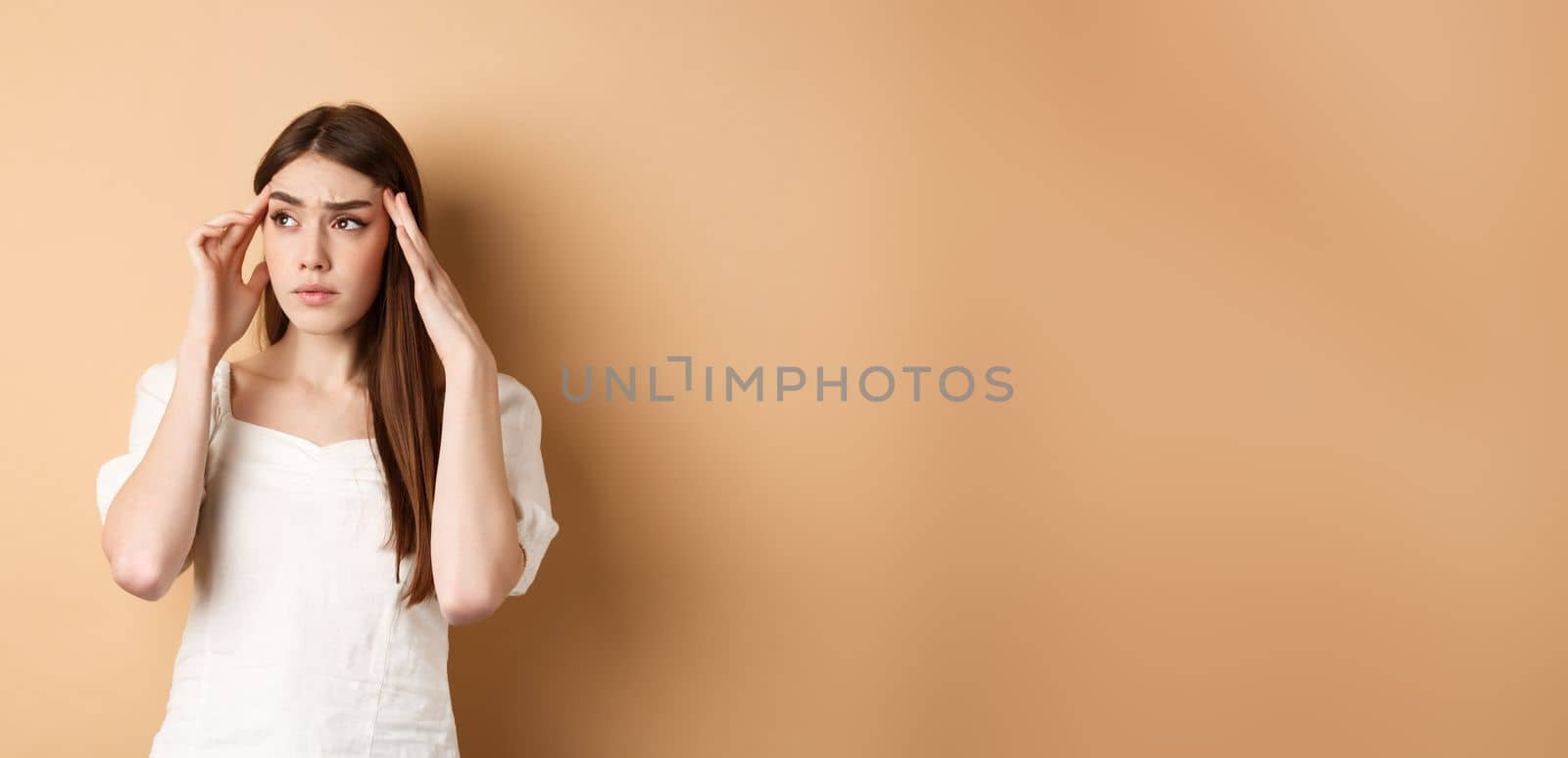 Troubled young woman feel sick, touching head temples, having headache, looking at upper left corner with tensed and distressed face, standing on beige background by Benzoix