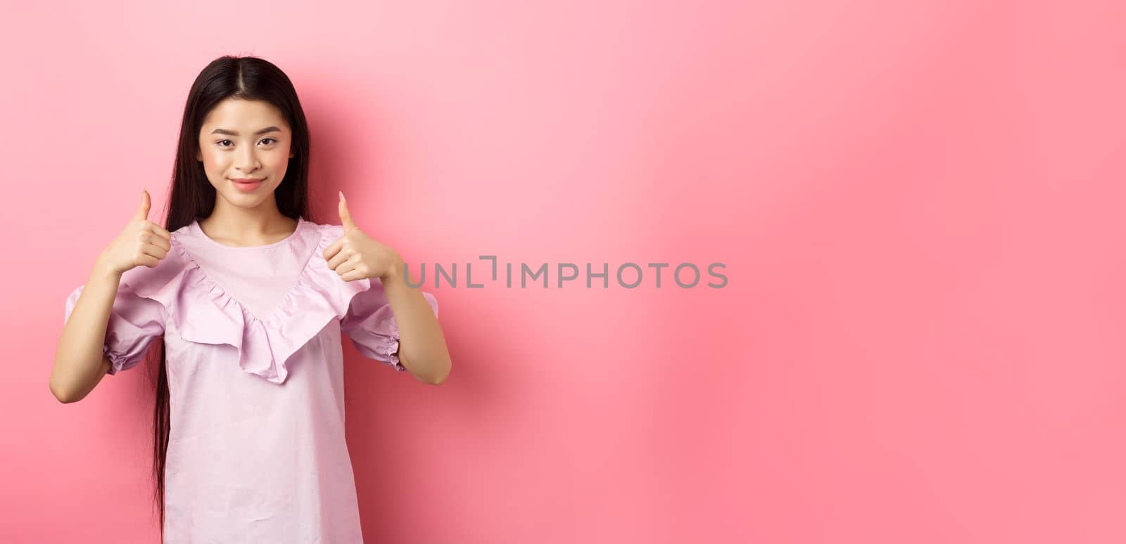 Well done. Smiling asian girl in dress showing thumbs up, like and approve product, praise great deal, standing against pink background by Benzoix