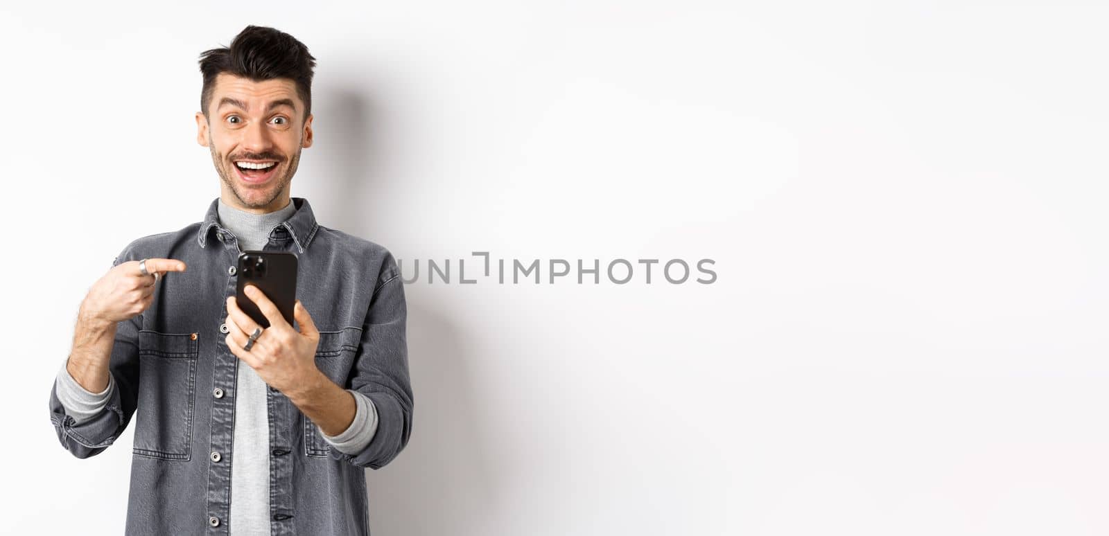 Excited young man receive good news on phone, pointing at smartphone and smiling happy, standing on white background.