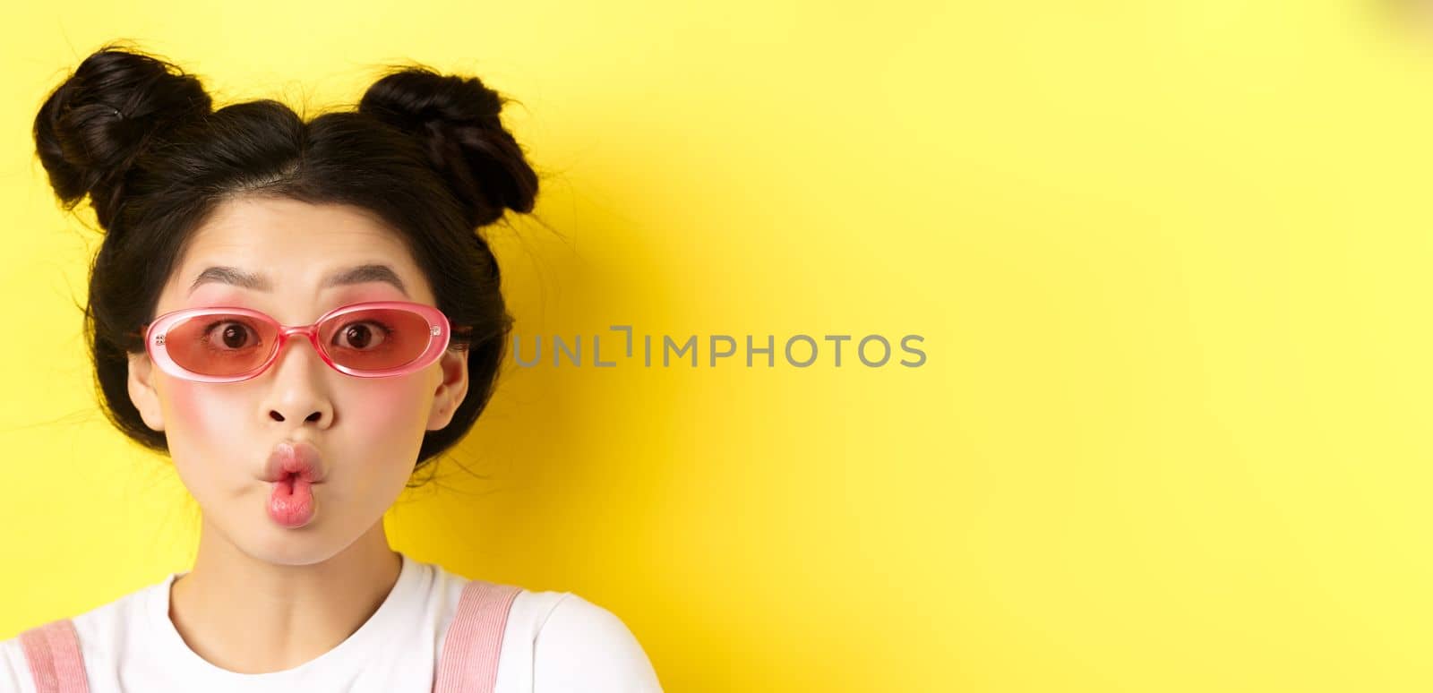 Summer fashion concept. Silly glamour girl in sunglasses, pucker lips like fish and looking funny at camera, standing against yellow background by Benzoix