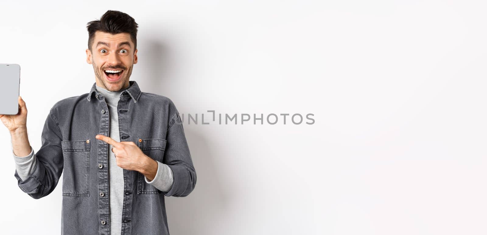 Excited handsome man showing empty smartphone screen and smiling, look here gesture, standing against white background.