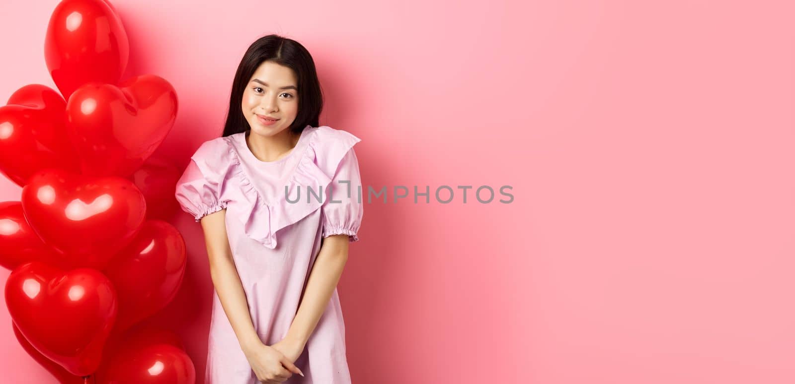 Cute asian girl in dress looking shy and smiling, standing modest near valentines day balloons, blushing on romantic date, looking at camera, pink background.