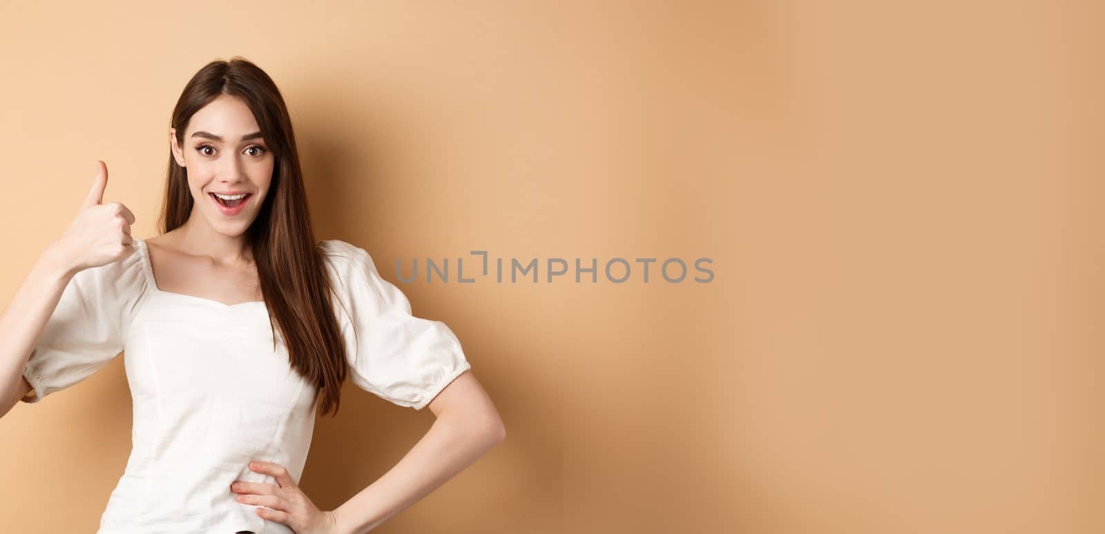 Amazed young woman showing thumb up, say yes and praise great product, approve choice, standing satisfied on beige background.