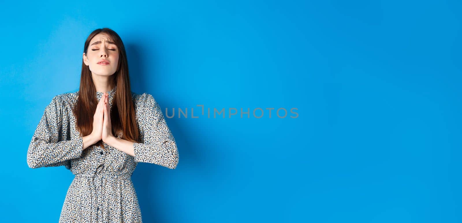 Pleading woman in dress begging for wish come true, supplicating god, standing against blue background. Copy space