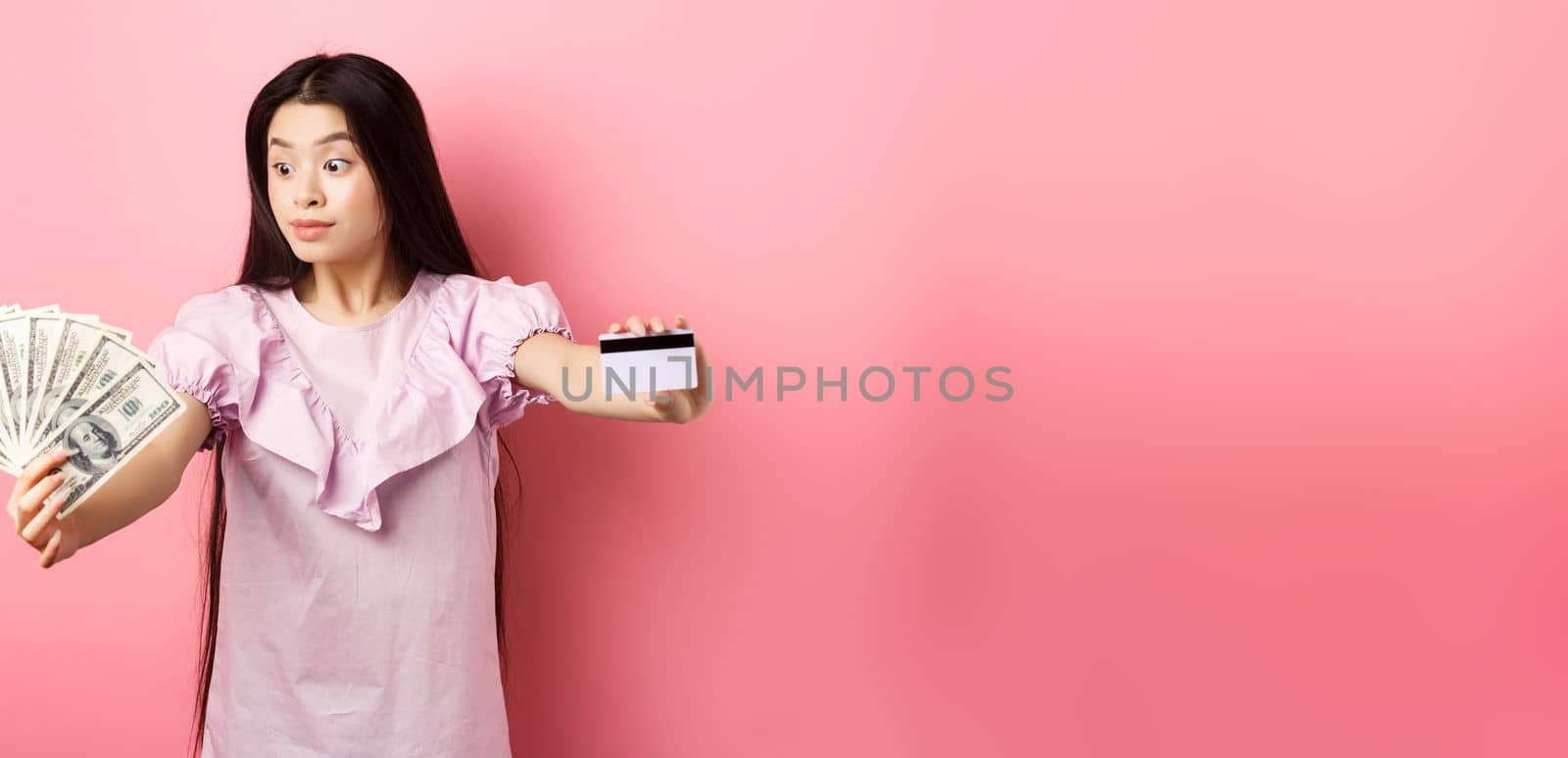 Excited teen asian girl stretch out hands with dollar bills and plastic credit card, look at money with amazement, standing on pink background by Benzoix