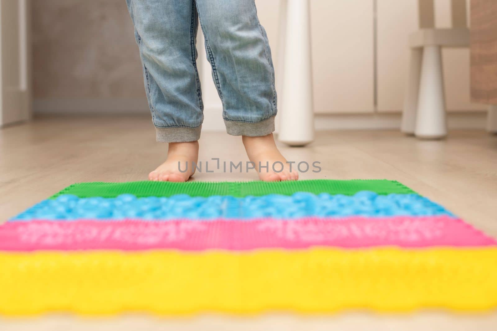 Health concept. A little boy stands with bare feet on a multi-colored orthopedic massage mat in a home interior. Close-up of children's heels. Back view.