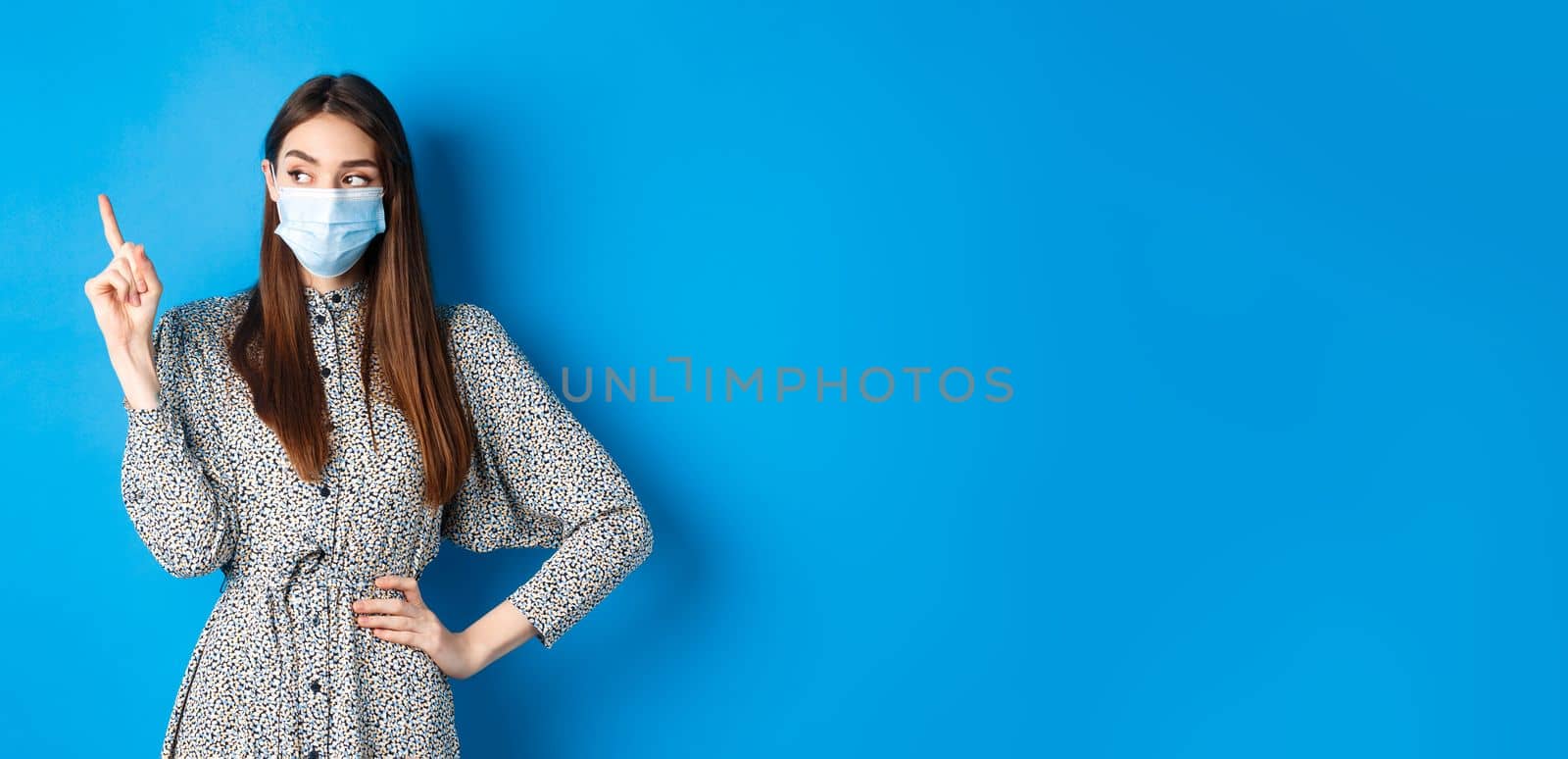 Healthy people and covid-19 pandemic concept. Curious girl in medical mask pointing at upper left corner, standing on blue background.