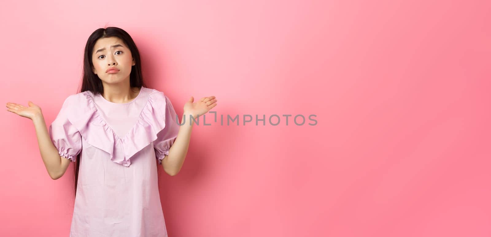 Sorry cant help. Clueless asian girl shrugging with sad face, know nothing, standing indecisive against pink background.