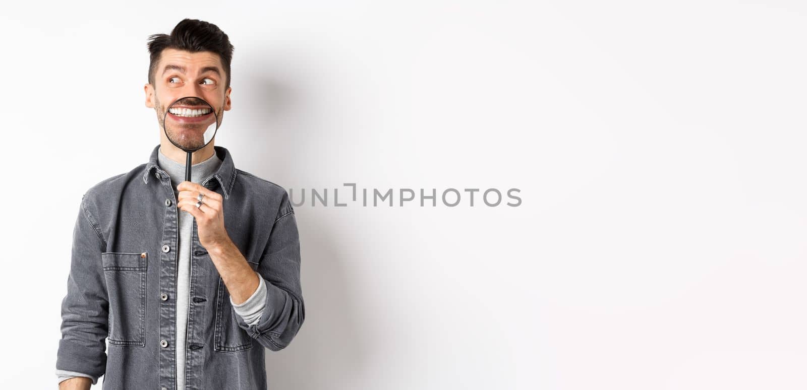 Handsome positive guy showing white perfect smile with magnifying glass, looking left at logo, standing against white background by Benzoix